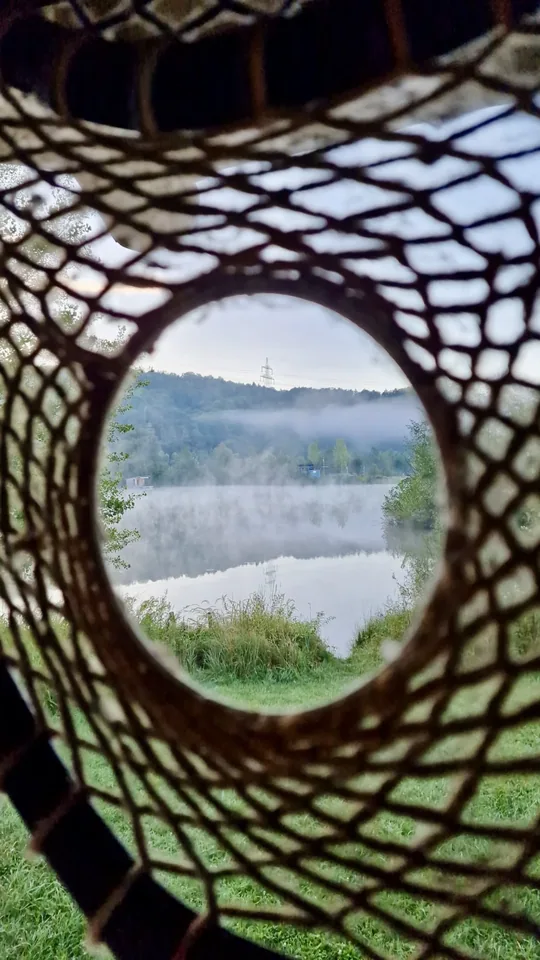 Blick durch einen geflochtenen Ring auf einen nebligen See und das grüne Ufer