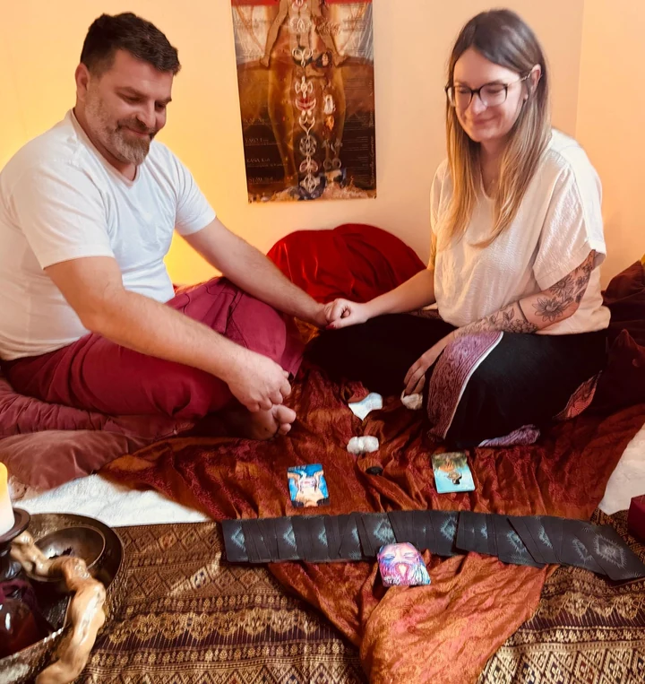 Ein Paar sitzt sich auf Kissen gegenüber und hält sich an den Händen, vor ihm liegen Karten auf einer Decke